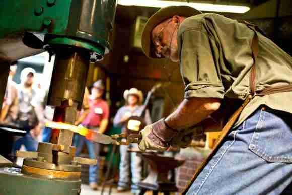 Blacksmith forging metal at a traditional workshop with onlookers in the background.