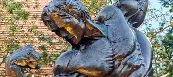Close-up of the bronze Arnold Schwarzenegger statue in Columbus, Ohio, showing the sculpted hair, downturned face, clenched fist and flexed upper body catching golden evening light, with trees behind.