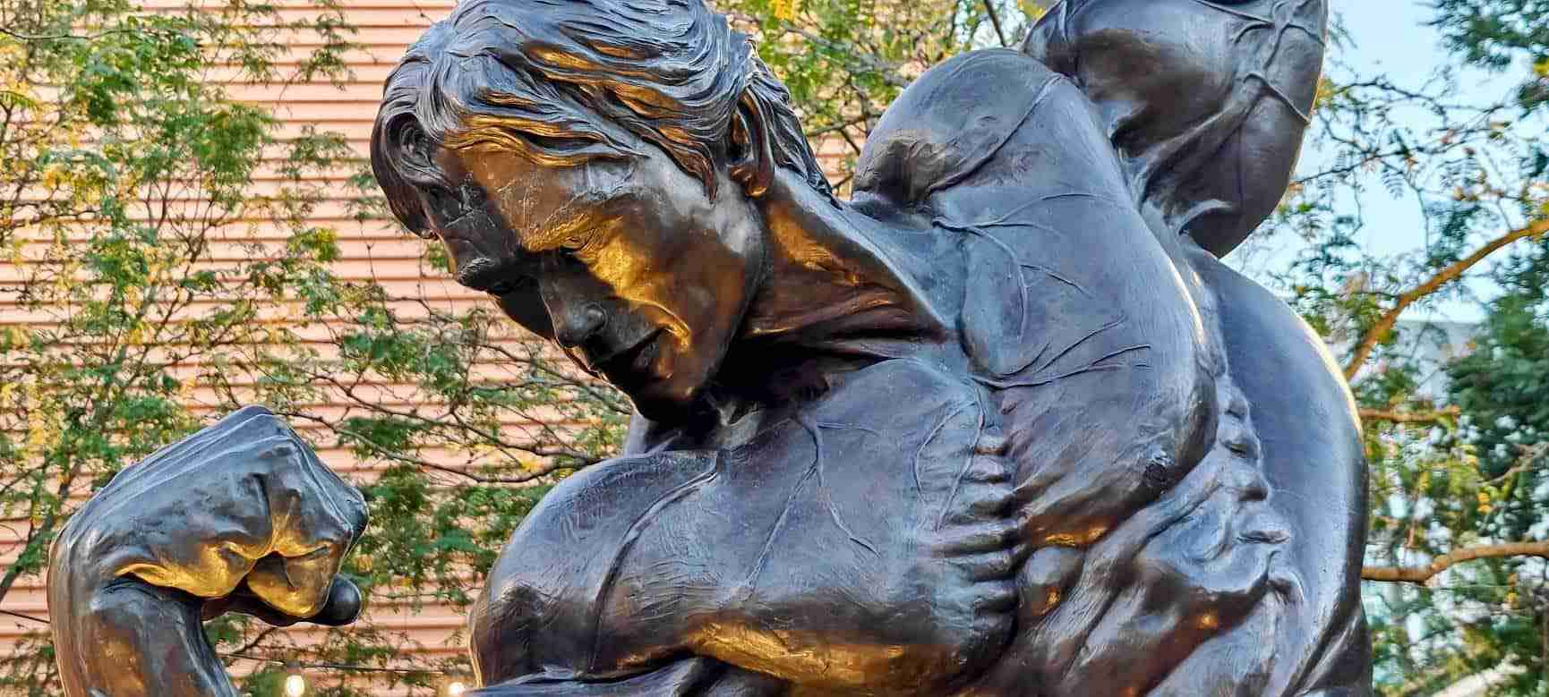 Close-up of the bronze Arnold Schwarzenegger statue in Columbus, Ohio, showing the sculpted hair, downturned face, clenched fist and flexed upper body catching golden evening light, with trees behind.